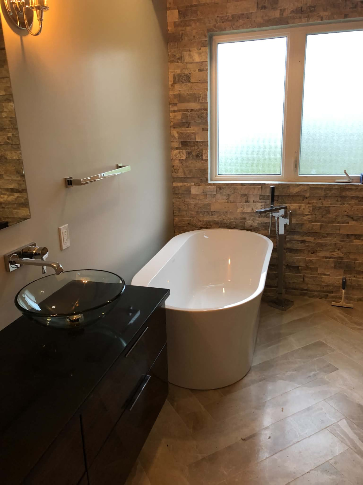 Freestanding white soaking tub with a chrome floor-mounted filler beside a clear glass vessel sink on a dark vanity, against a stacked stone feature wall