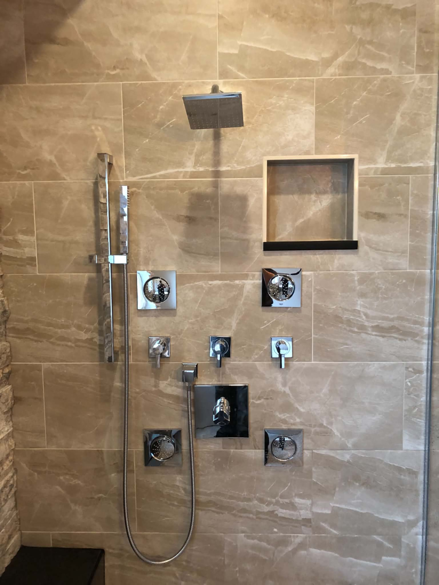 Walk-in shower with a square overhead rain showerhead, a hand wand on a slide bar, four body sprays and three chrome controls set into cream marble-patterned tile
